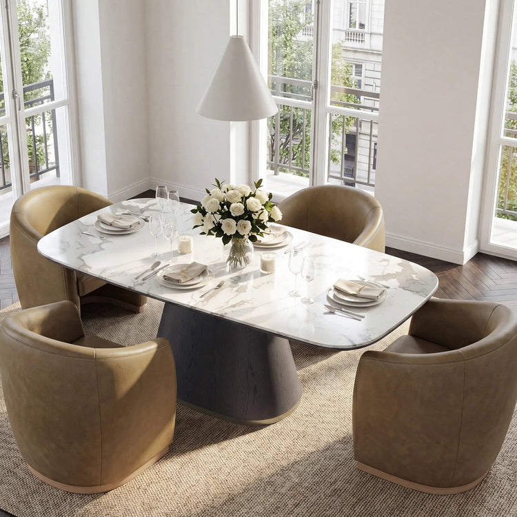 A modern dining room scene with a white marble-top dining table on a dark pedestal base, surrounded by four taupe upholstered armchairs. The table is set with plates, glasses, and a white rose centerpiece, with large windows and a white pendant lamp in the background.