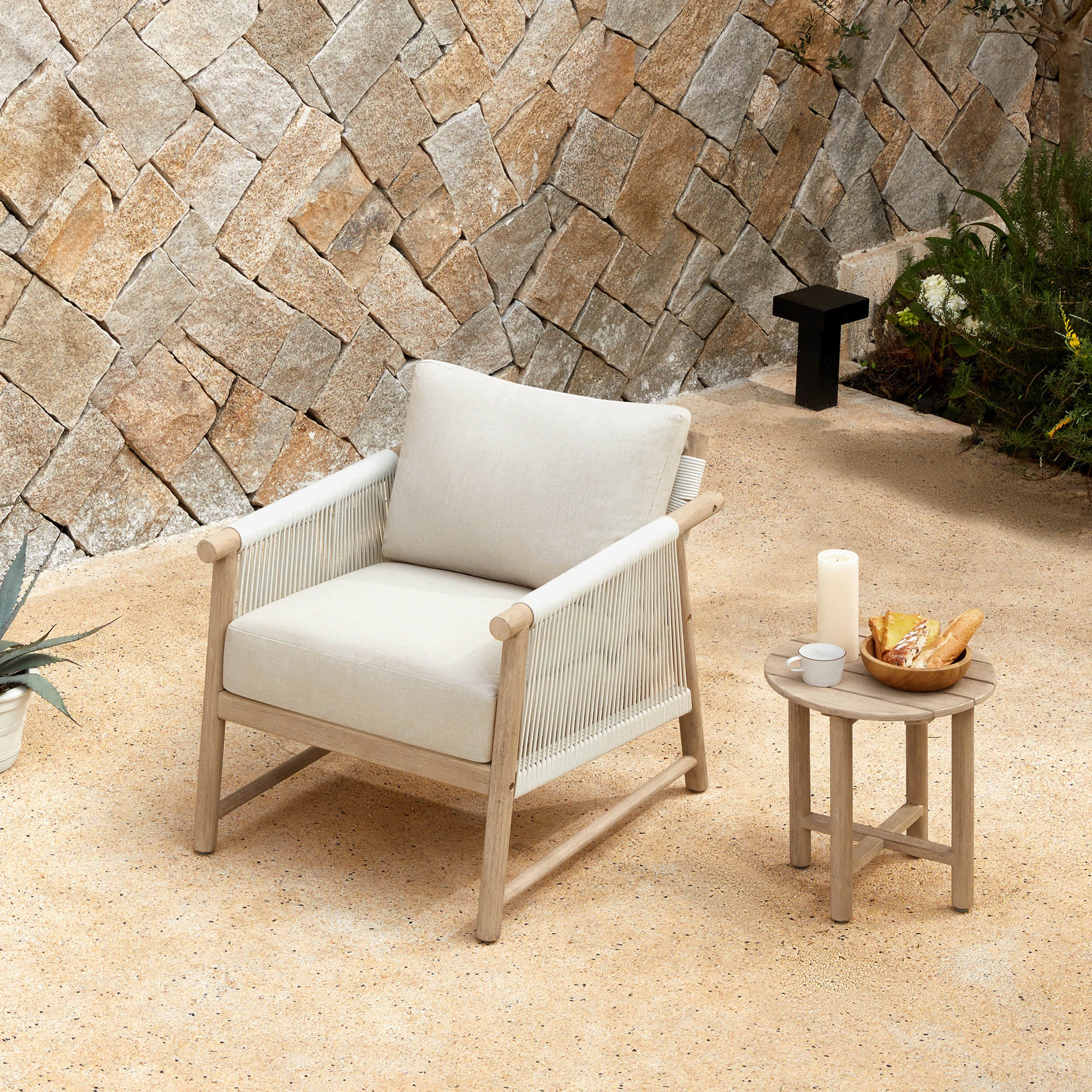 A Sienna Modern Acacia wood woven outdoor lounge chair with a cushion, beside a small table with a candle and bread.