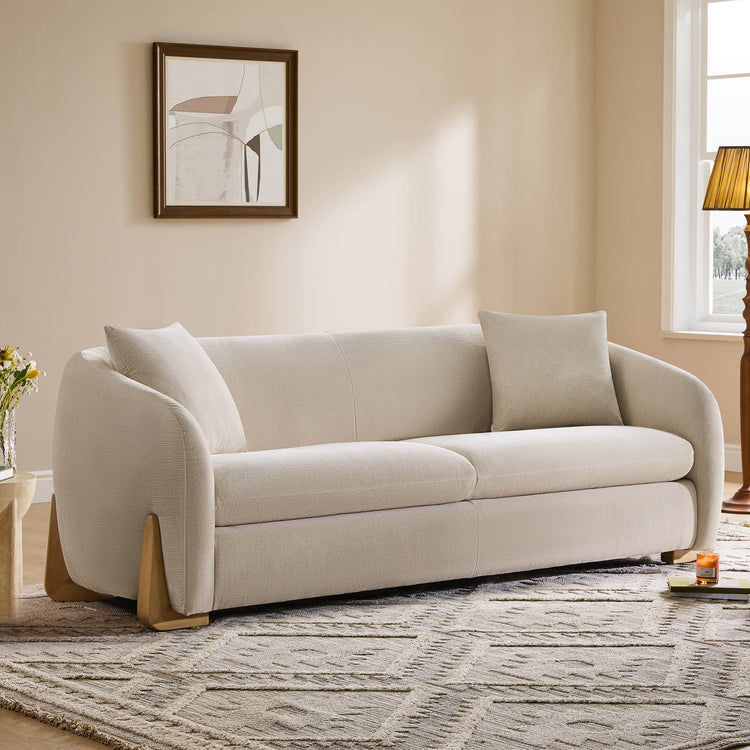 A cozy cream sofa with rounded armrests and wooden legs, set in a warmly lit living room with abstract art on the wall and a patterned rug on the floor.