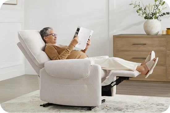 An elderly woman reclining in a white armchair, reading a magazine in a cozy room.