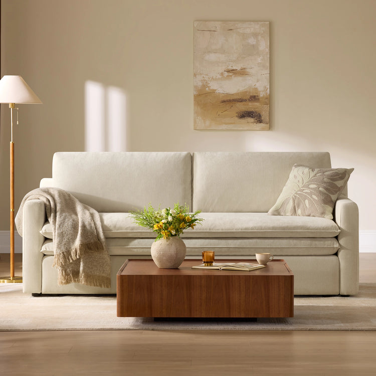A cream-colored upholstered sofa in a bright, modern living room setting, styled with a beige throw blanket, a patterned pillow, a wooden coffee table, and a floor lamp.