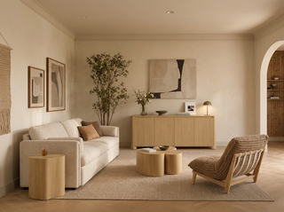 A warm neutral living room with a light oak sideboard against the back wall, a cream sofa, round oak coffee tables, a sculptural wooden lounge chair, abstract artwork, and a tall potted tree.