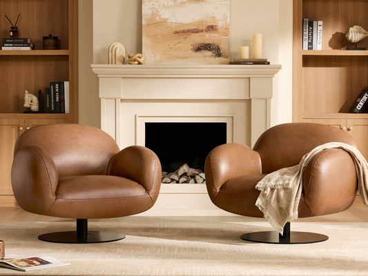 Pair of modern cognac brown leather swivel armchairs placed in front of a white fireplace mantle and tall wooden bookshelves.