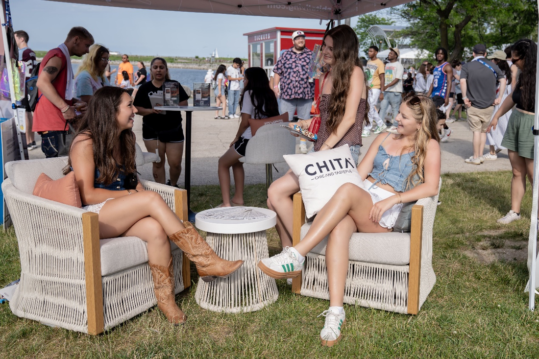 Two women sit on stylish chairs, chatting and enjoying a sunny outdoor event by the water.