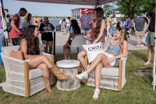 Two women sit on stylish chairs, chatting and enjoying a sunny outdoor event by the water.