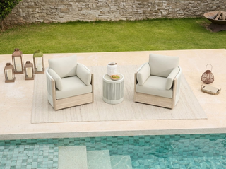 Two cream-cushioned outdoor patio club chairs with a round side table sit poolside on a beige rug, illustrating coordinated outdoor furniture styling.