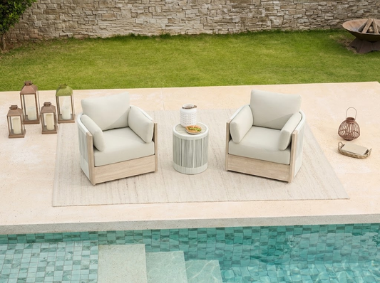 Two cream-cushioned outdoor patio club chairs with a round side table sit poolside on a beige rug, illustrating coordinated outdoor furniture styling.