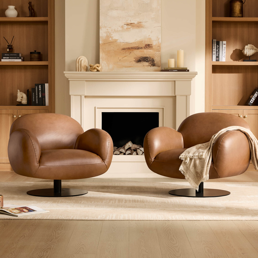 Pair of modern cognac brown leather swivel armchairs placed in front of a white fireplace mantle and tall wooden bookshelves.