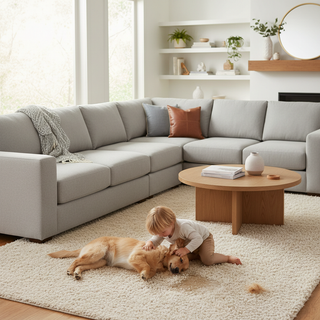A bright and airy living room featuring a modern modular sectional sofa in a neutral-toned performance fabric. A child and a dog play on a rug nearby where a small spill and some fur are visible, while the sofa remains pristine and clean.