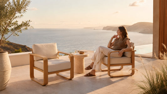 Two modern patio armchairs with cream cushions on a seaside terrace, with a woman relaxing beside a round side table at sunset.
