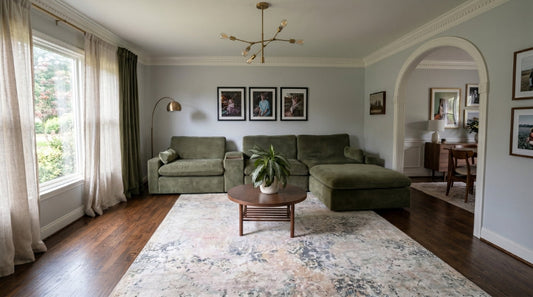 Green modular sectional sofa surrounds a round wooden coffee table with a leafy plant, framed photos, and patterned rug in a living room.