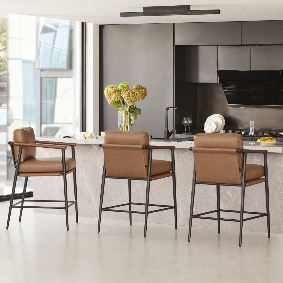 Three tan leather counter stools with black metal frames line a marble kitchen island in a bright modern kitchen with dark cabinetry and flowers