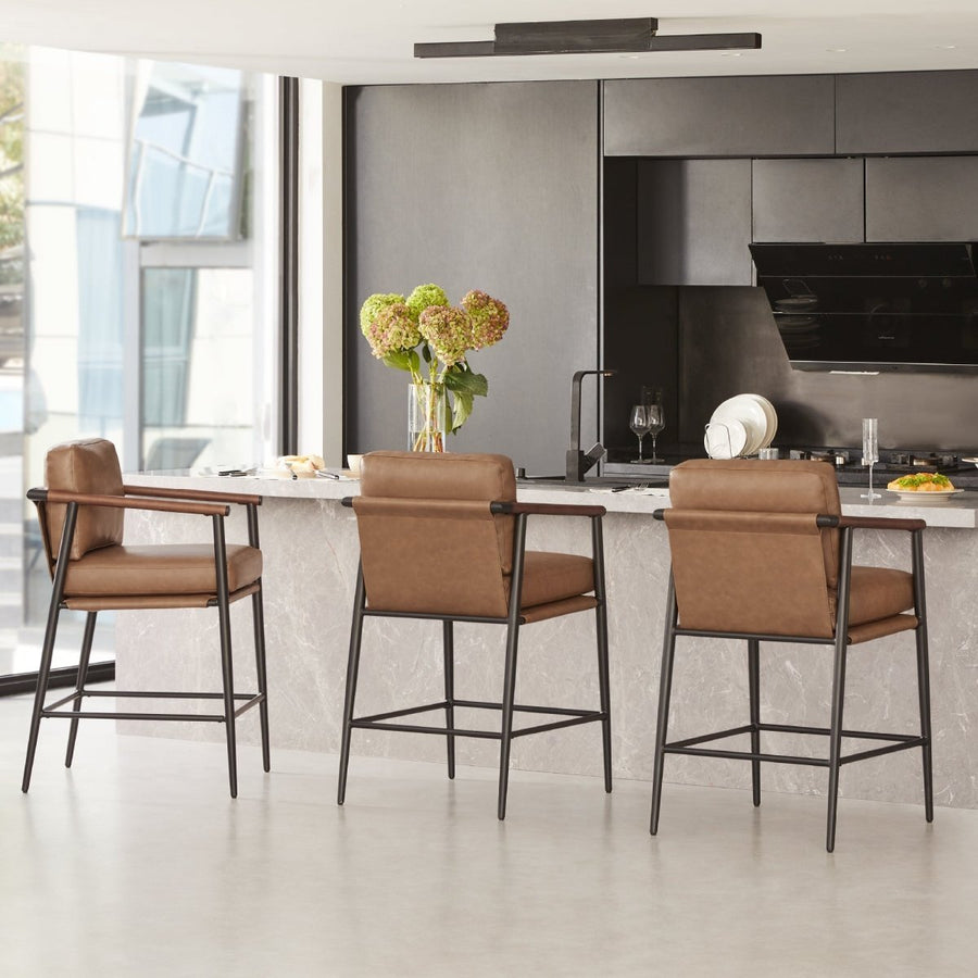Three tan leather counter stools with black metal frames line a marble kitchen island in a bright modern kitchen with dark cabinetry and flowers