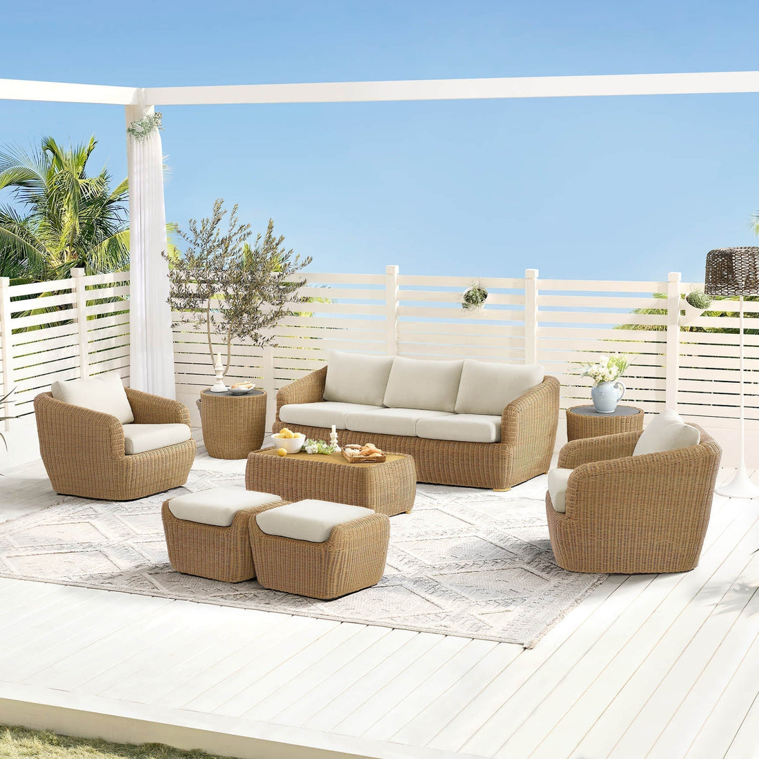 Light wicker outdoor sofa set with cream cushions and ottoman arranged on a sunny patio near white fencing.