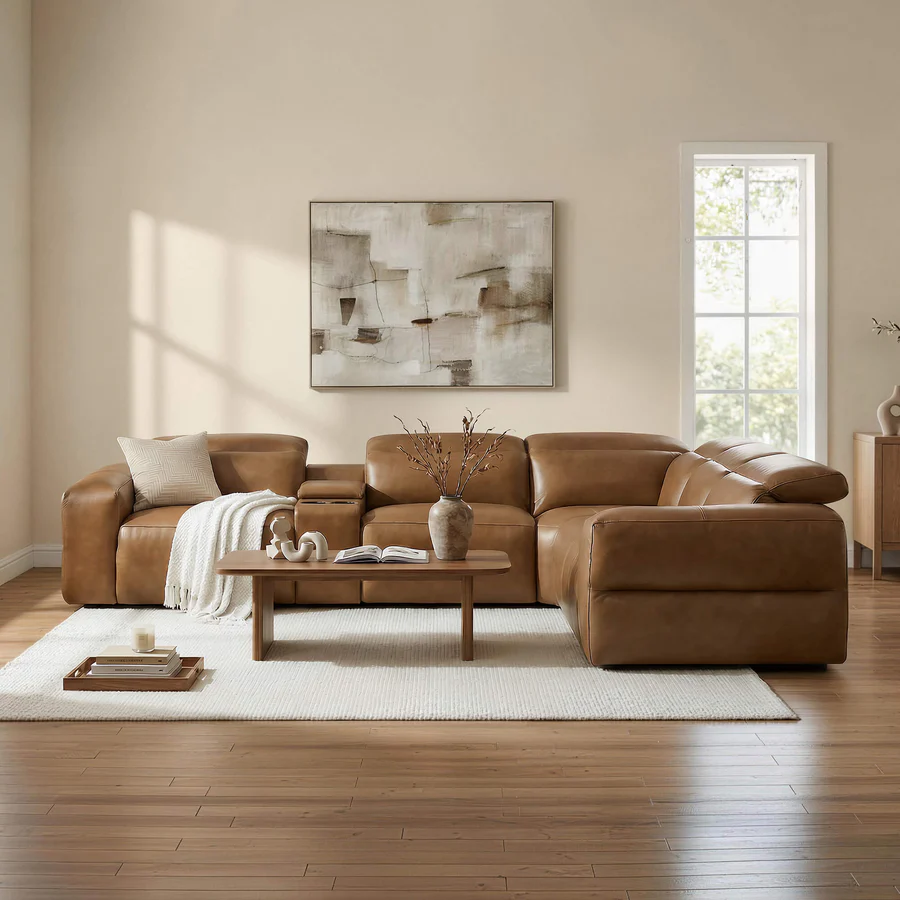 Warm neutral living room with a caramel leather deep-seat sectional, white rug, wood coffee table, abstract wall art, and sunlight through tall windows.