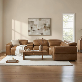 Warm neutral living room with a caramel leather deep-seat sectional, white rug, wood coffee table, abstract wall art, and sunlight through tall windows.