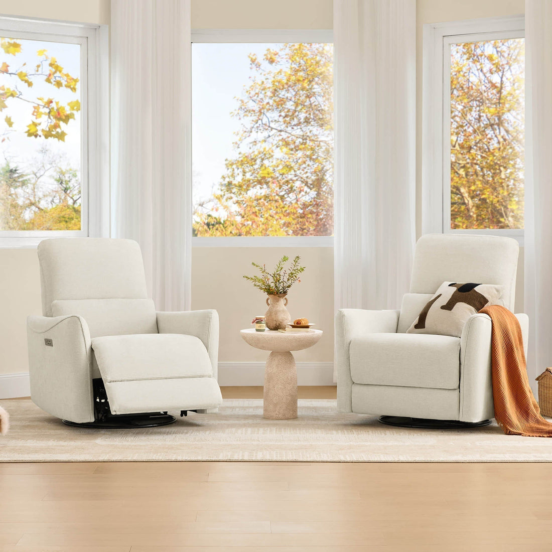 Two cream recliner chairs sit beside a round table in a bright living room with large windows and autumn trees outside.