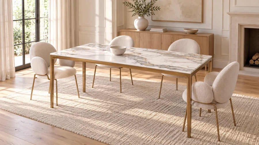 White veined rectangular dining table with slender brass-colored legs and beige chairs in a bright room.