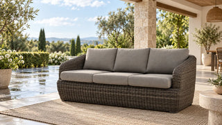 A grey-cushioned Liana wicker sofa on a covered terrace beside rain-damp paving