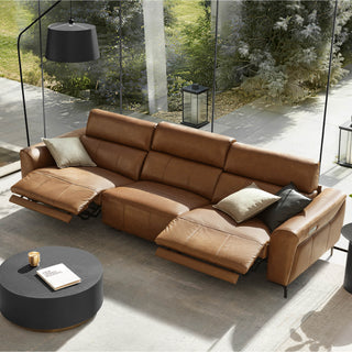 A modern brown leather power reclining sofa with open footrests, a black round coffee table, and large glass walls looking onto a garden.