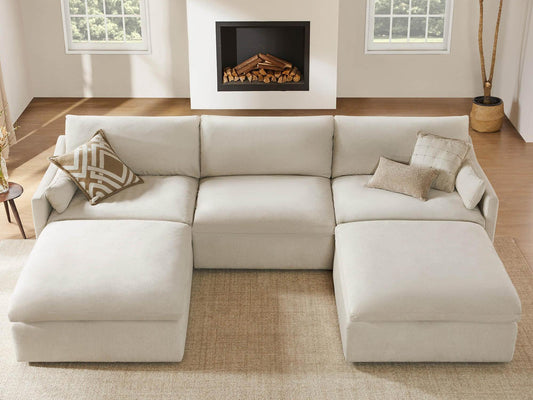 Cream performance fabric modular sectional sofa with two chaise lounges in a sunlit living room with wood floors, fireplace, large windows, neutral pillows, and a potted tree.