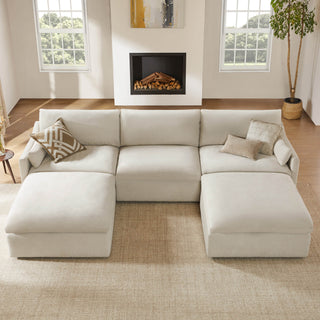 Cream performance fabric modular sectional sofa with two chaise lounges in a sunlit living room with wood floors, fireplace, large windows, neutral pillows, and a potted tree.