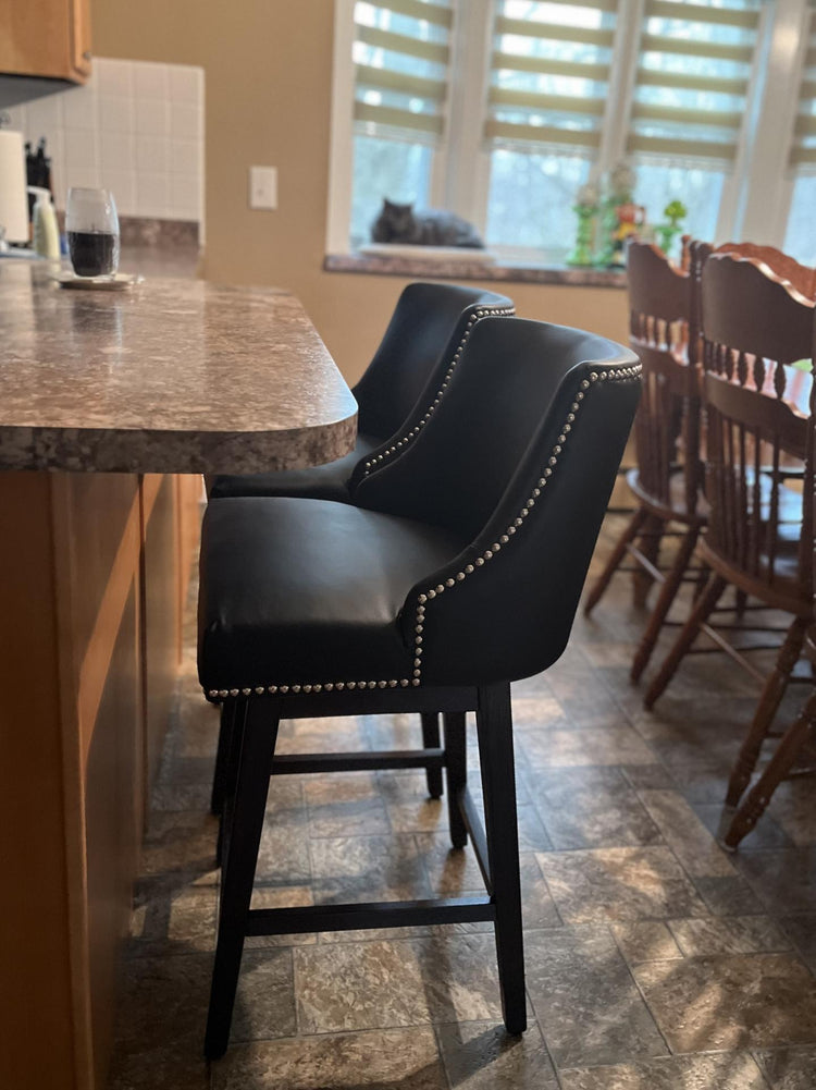 Dark upholstered counter stool with nailhead trim placed beside a kitchen island, shown in a home dining area with wood bar chairs nearby and window.