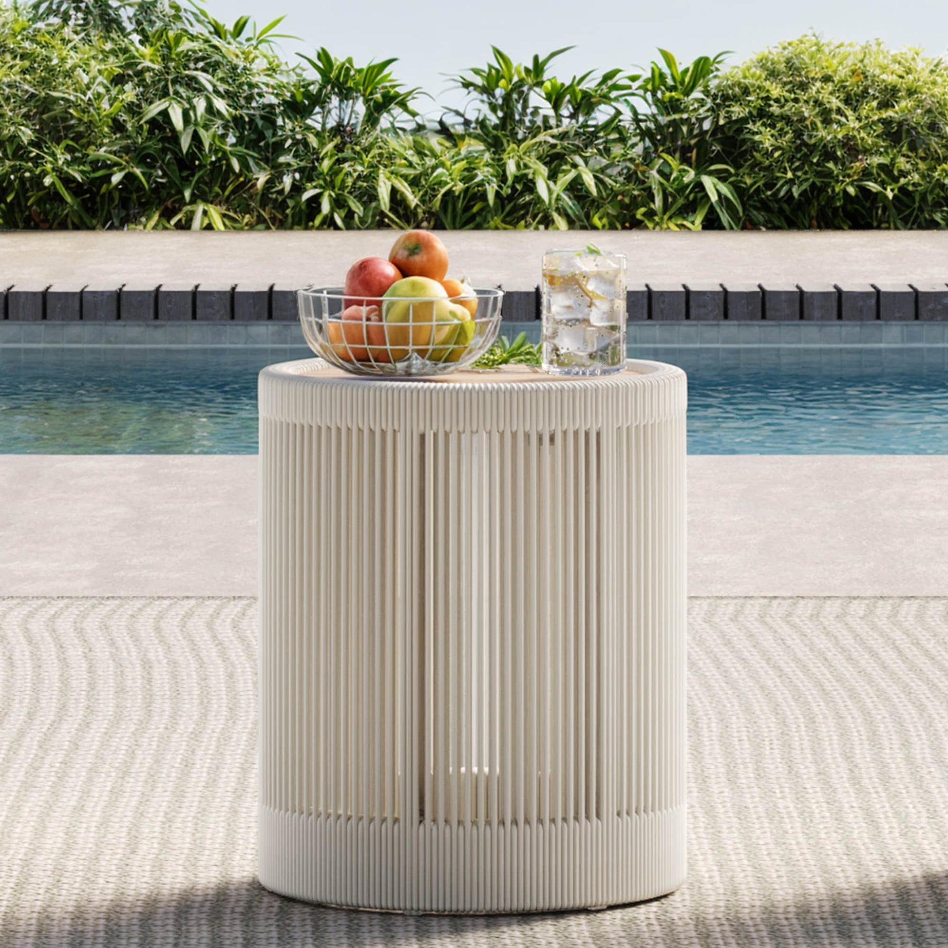 A Clover Acacia Wood Woven Outdoor Bistro Set side table with fruits and a drink by the pool.