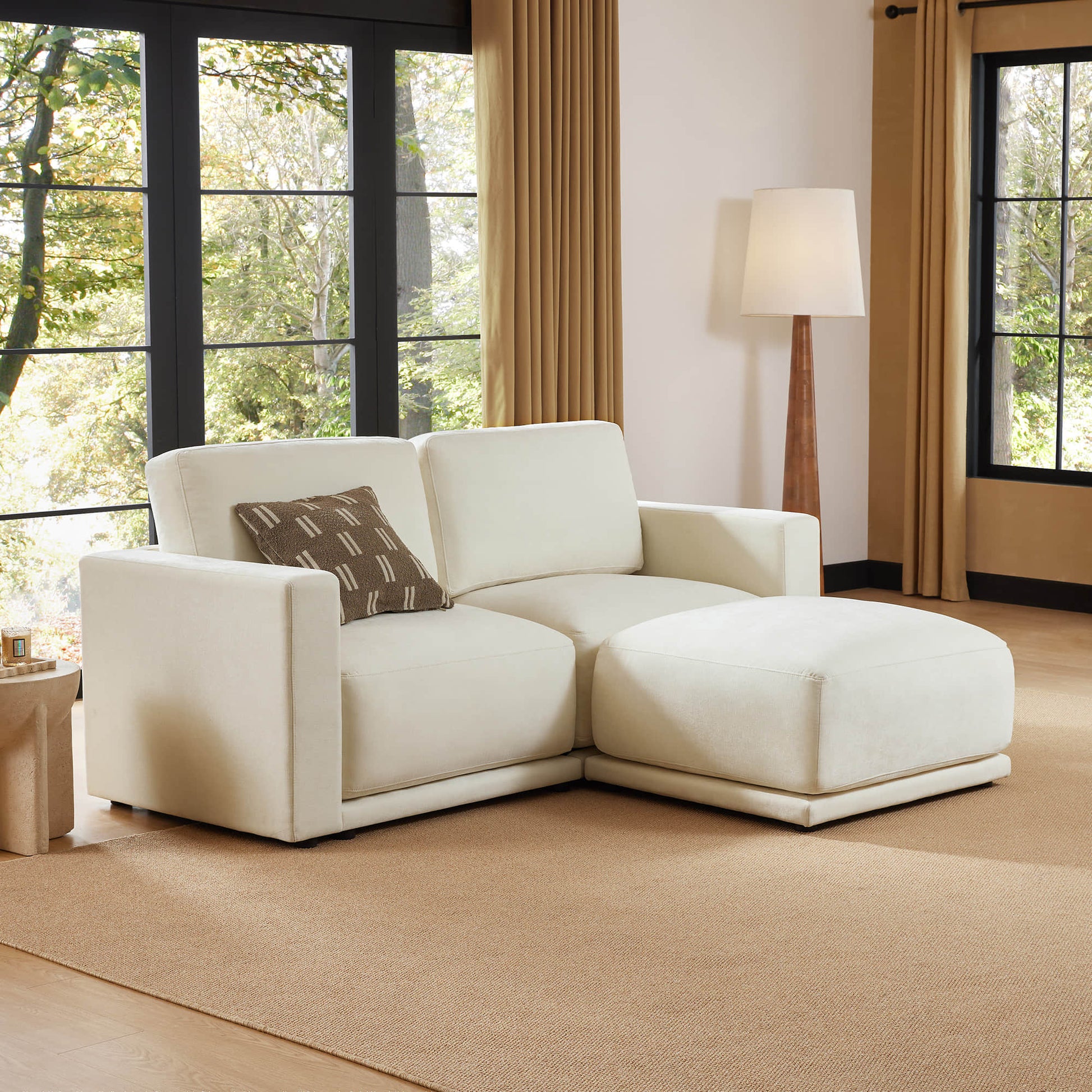 Beige sectional sofa with an ottoman in a living room featuring large windows, a floor lamp, and neutral décor. The space is bright and welcoming, with a cozy atmosphere.