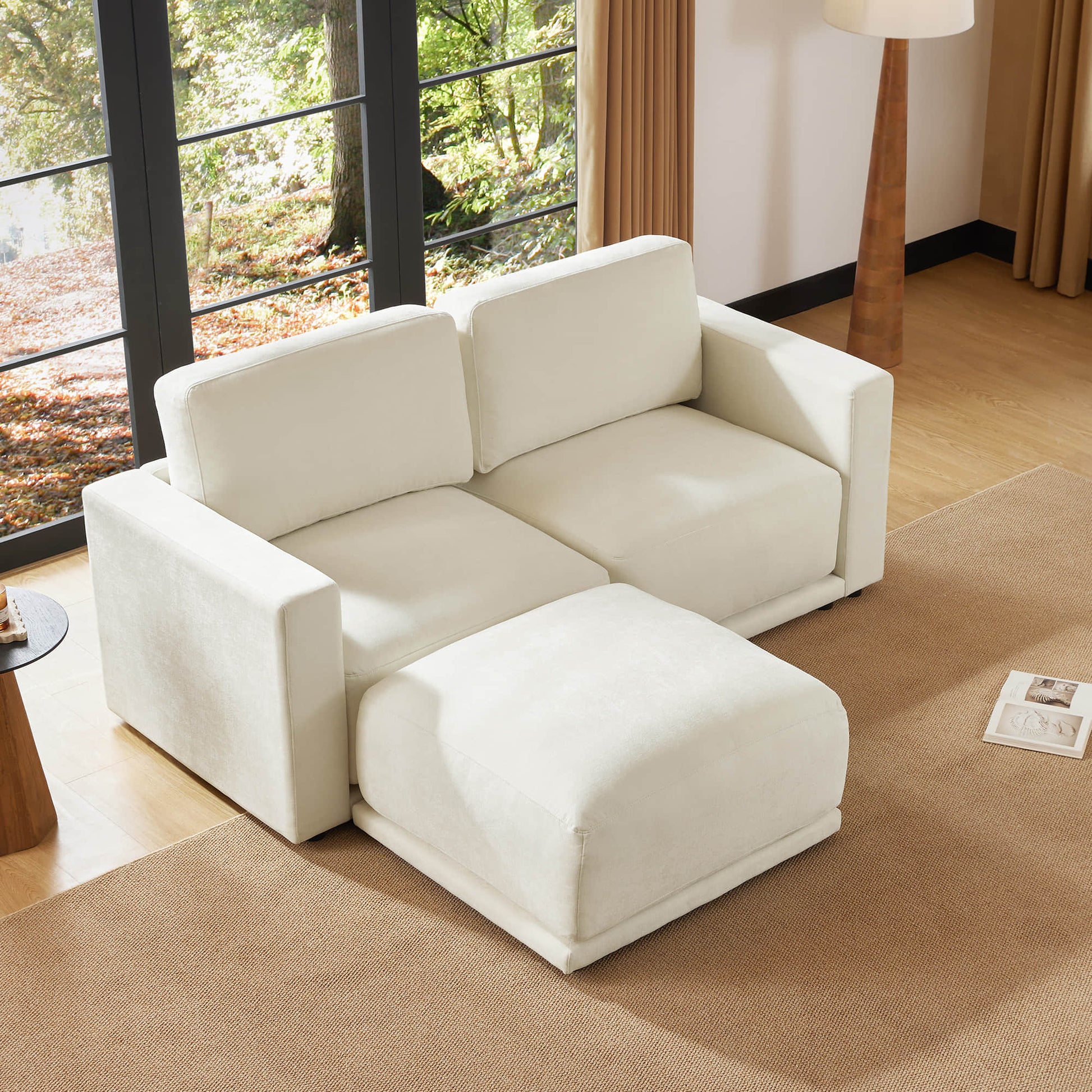 Beige sectional sofa with an ottoman in a bright living room with large windows, a floor lamp, and a neutral-toned rug. The room is filled with natural light and has a cozy, inviting atmosphere.
