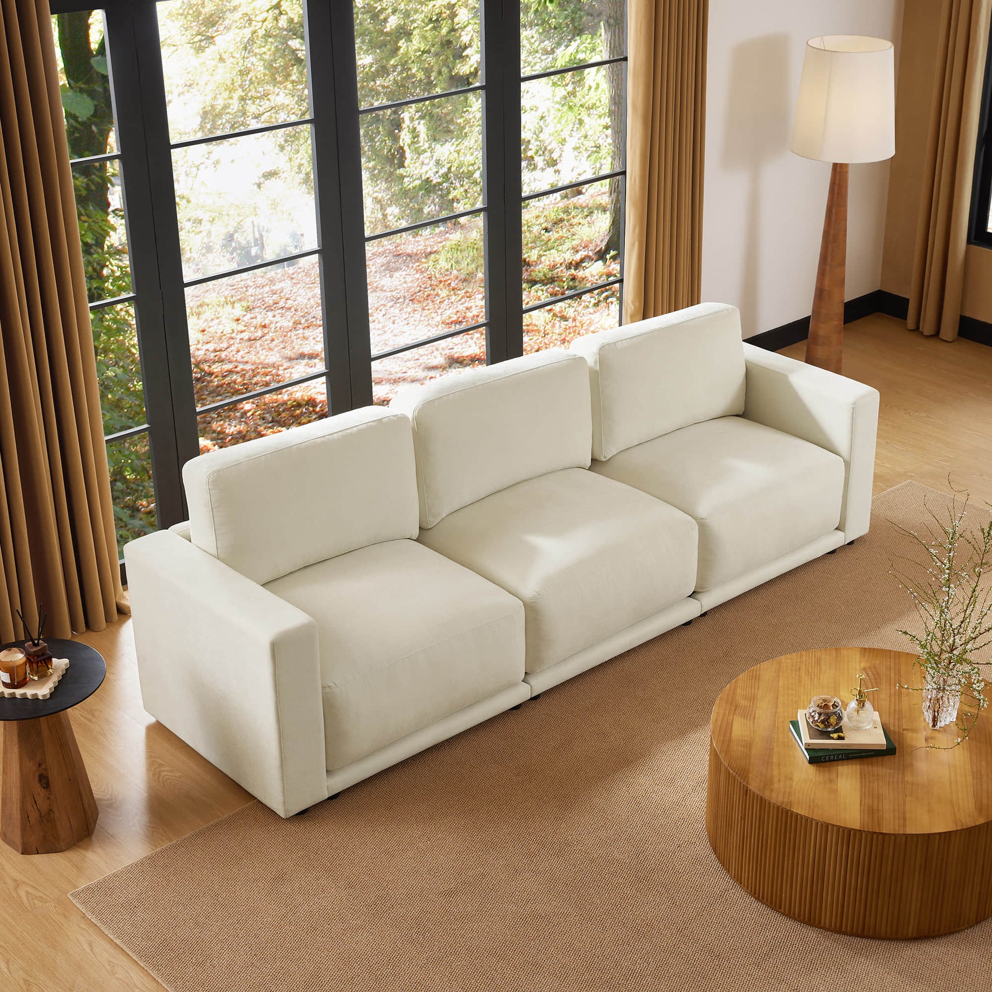 Top view of a beige sectional sofa in a living room with large windows, a round wooden coffee table, and a floor lamp. The room is decorated with neutral tones and natural light.