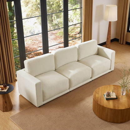 Top view of a beige sectional sofa in a living room with large windows, a round wooden coffee table, and a floor lamp. The room is decorated with neutral tones and natural light.