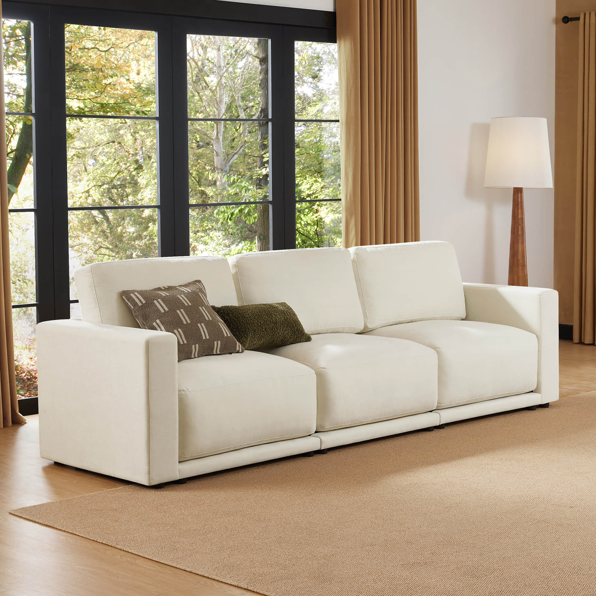 Front view of a beige modular sectional sofa in a living room setting, with decorative pillows and large windows in the background.