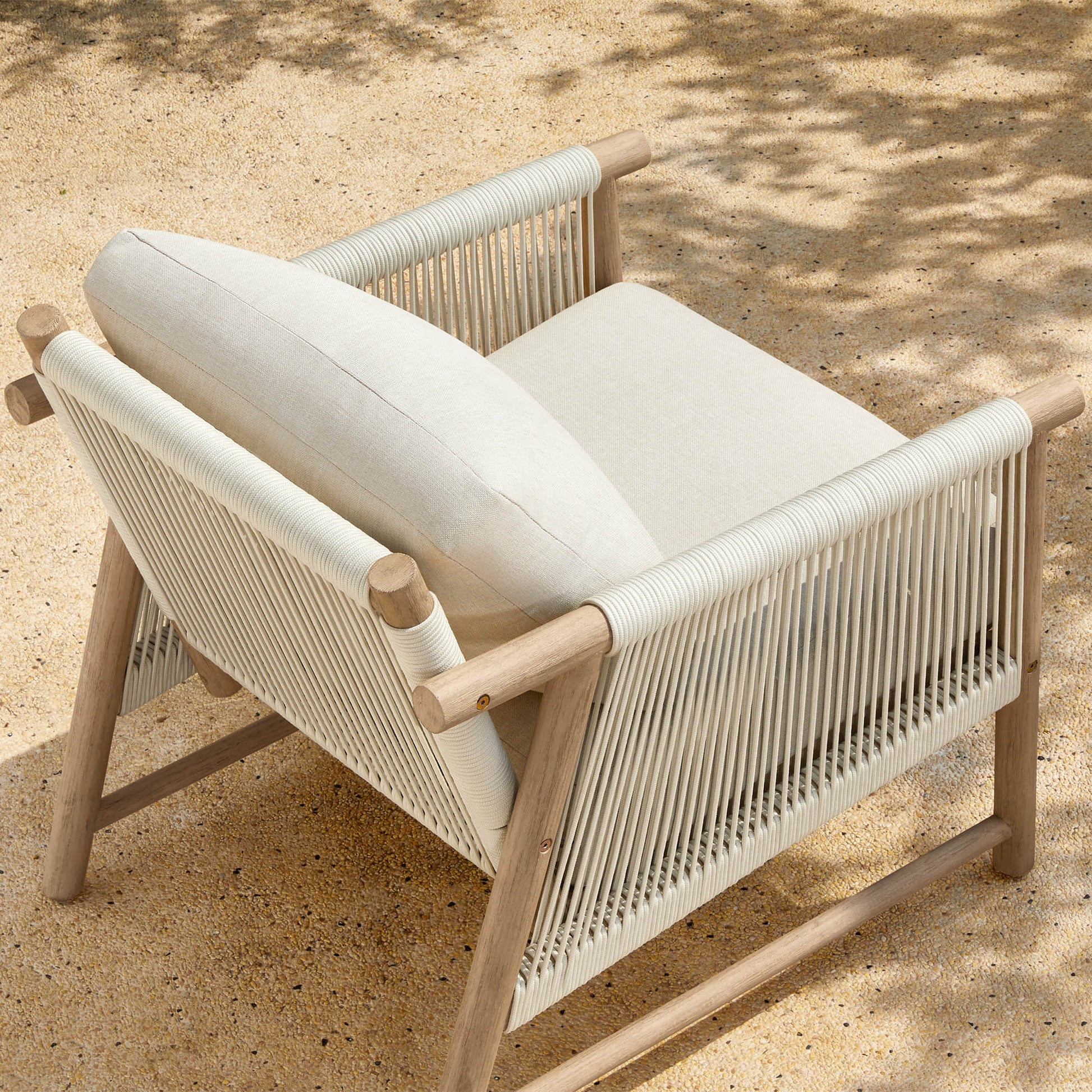 A Sienna Modern Acacia Wood Woven Outdoor Bistro Set chair with light beige cushions on a sandy surface.