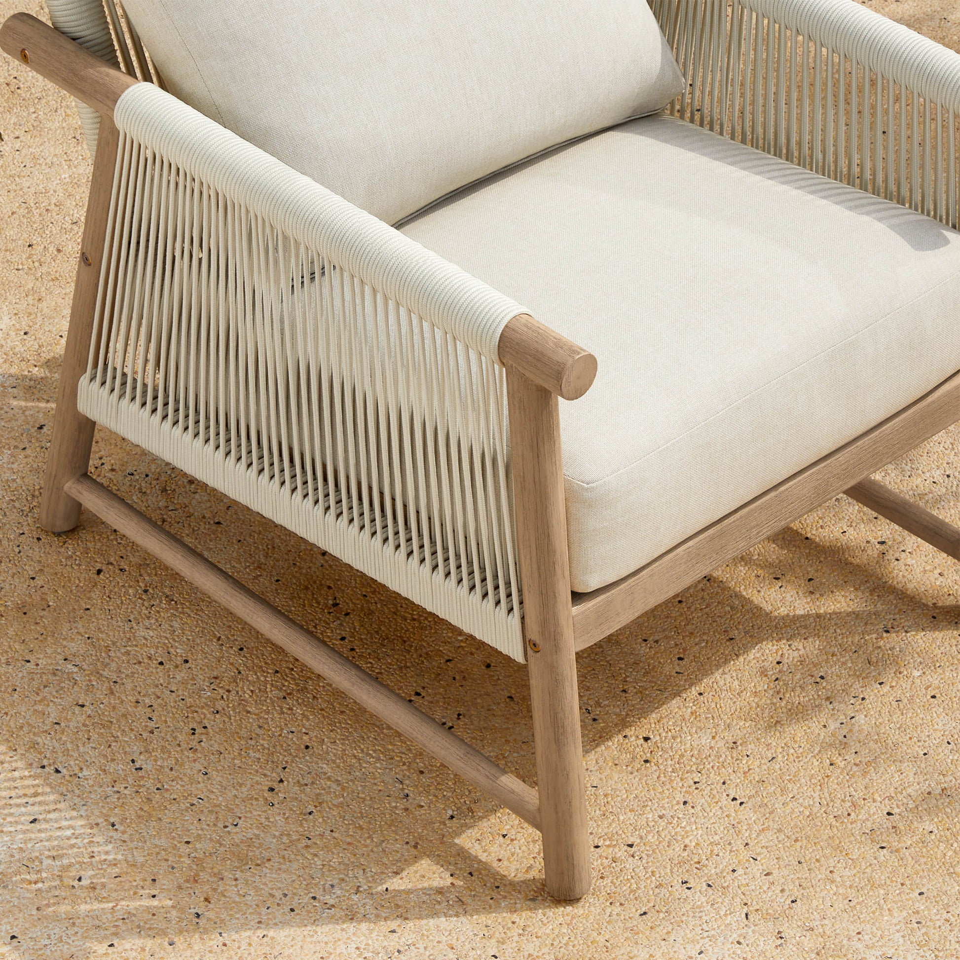 A Sienna Modern Acacia Wood Woven Outdoor Bistro Set chair with light beige cushions on a sandy surface.