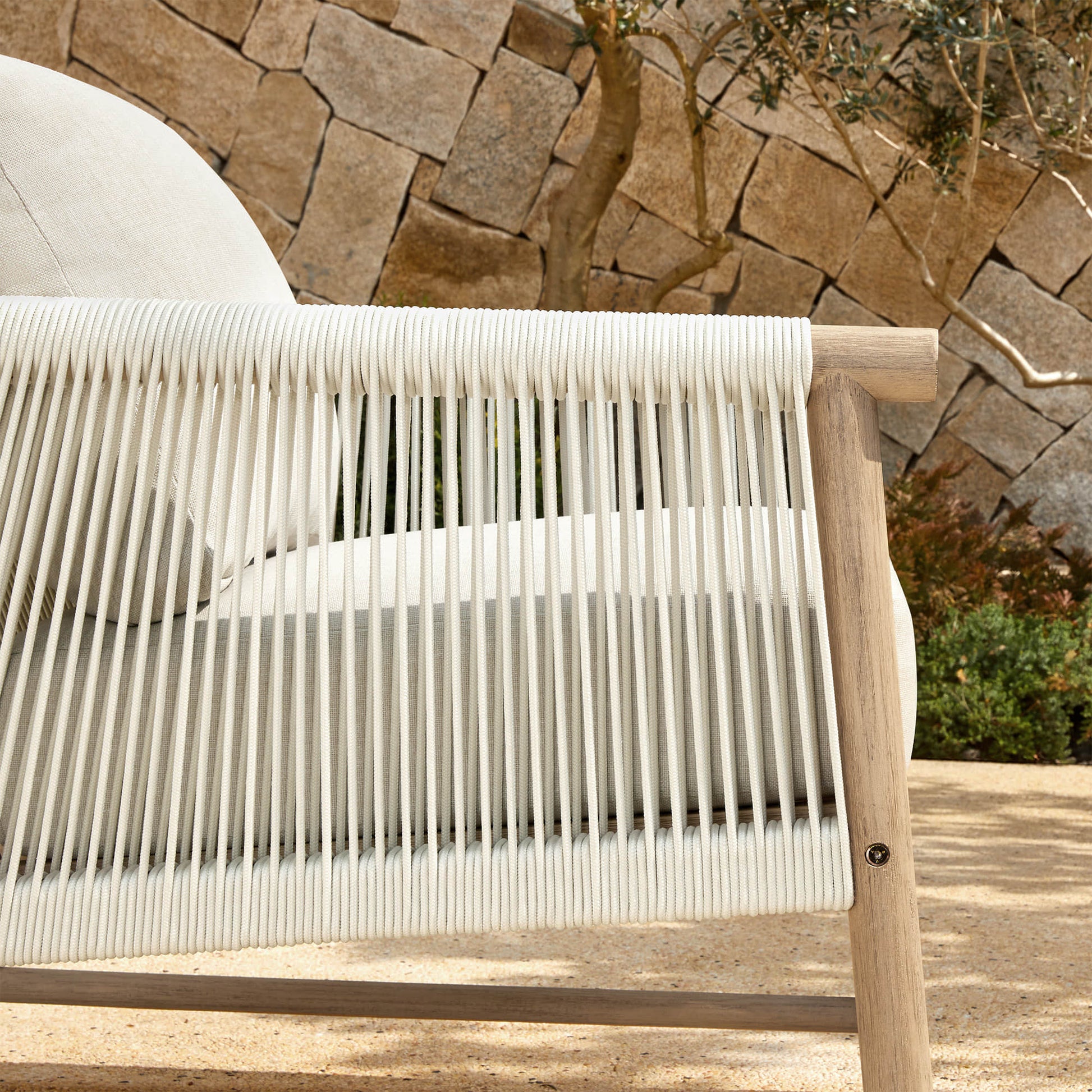 A Sienna Modern Acacia Wood Woven Outdoor Bistro Set chair with light beige cushions, set against a stone wall.