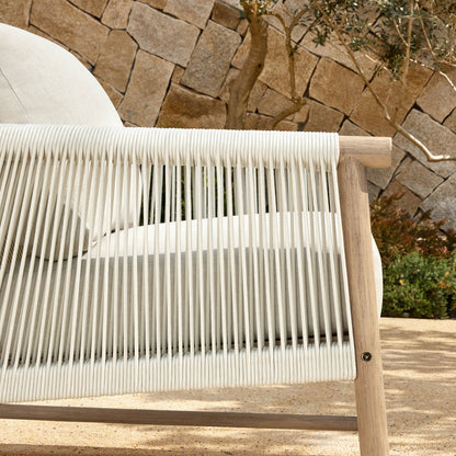 A Sienna Modern Acacia Wood Woven Outdoor Bistro Set chair with light beige cushions, set against a stone wall.