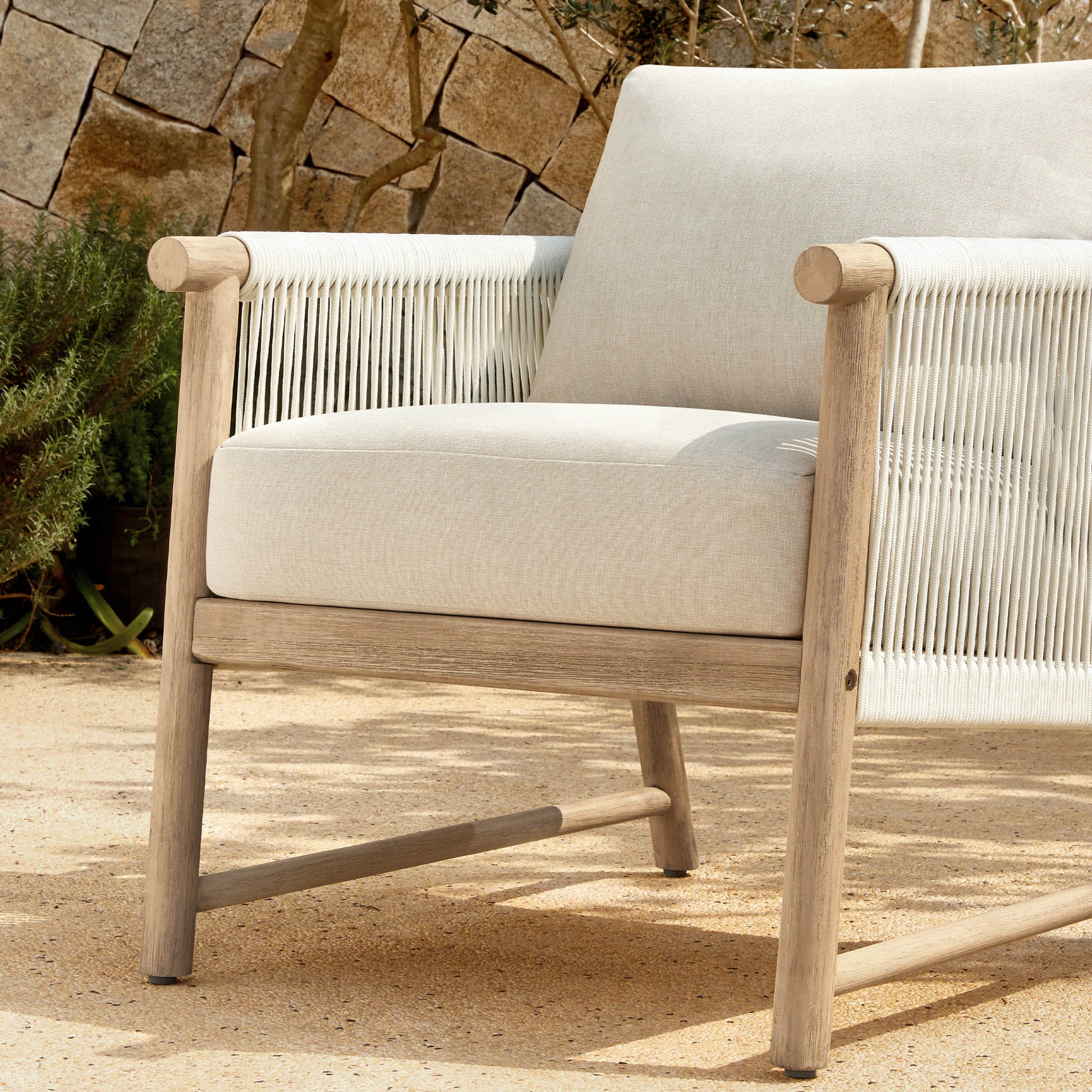 A Sienna Modern Acacia Wood Woven Outdoor Bistro Set chair with light beige cushions, placed on a sandy patio.