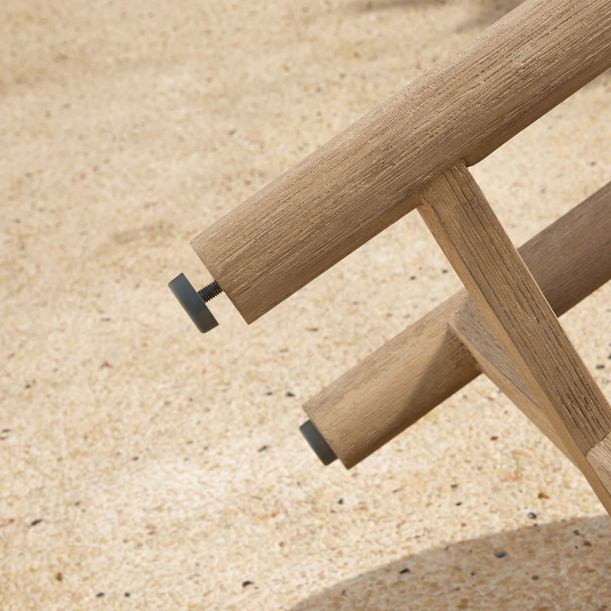 A close-up of a Sienna Modern Acacia Wood Woven Outdoor Bistro Set chair leg with black caps, on a sandy surface.