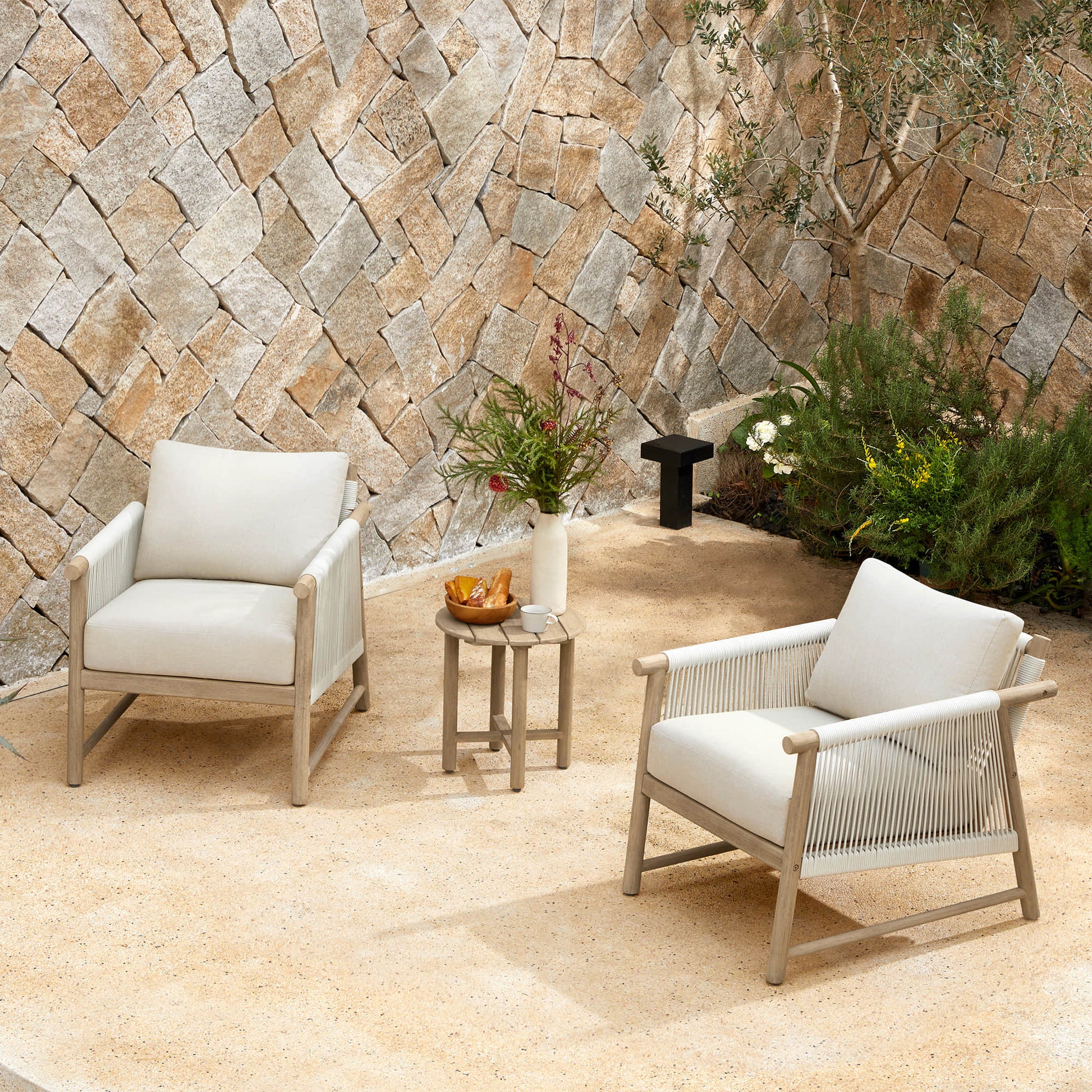 Two Sienna Modern Acacia Wood Woven Outdoor Bistro Set chairs with light beige cushions on a patio near a stone wall.
