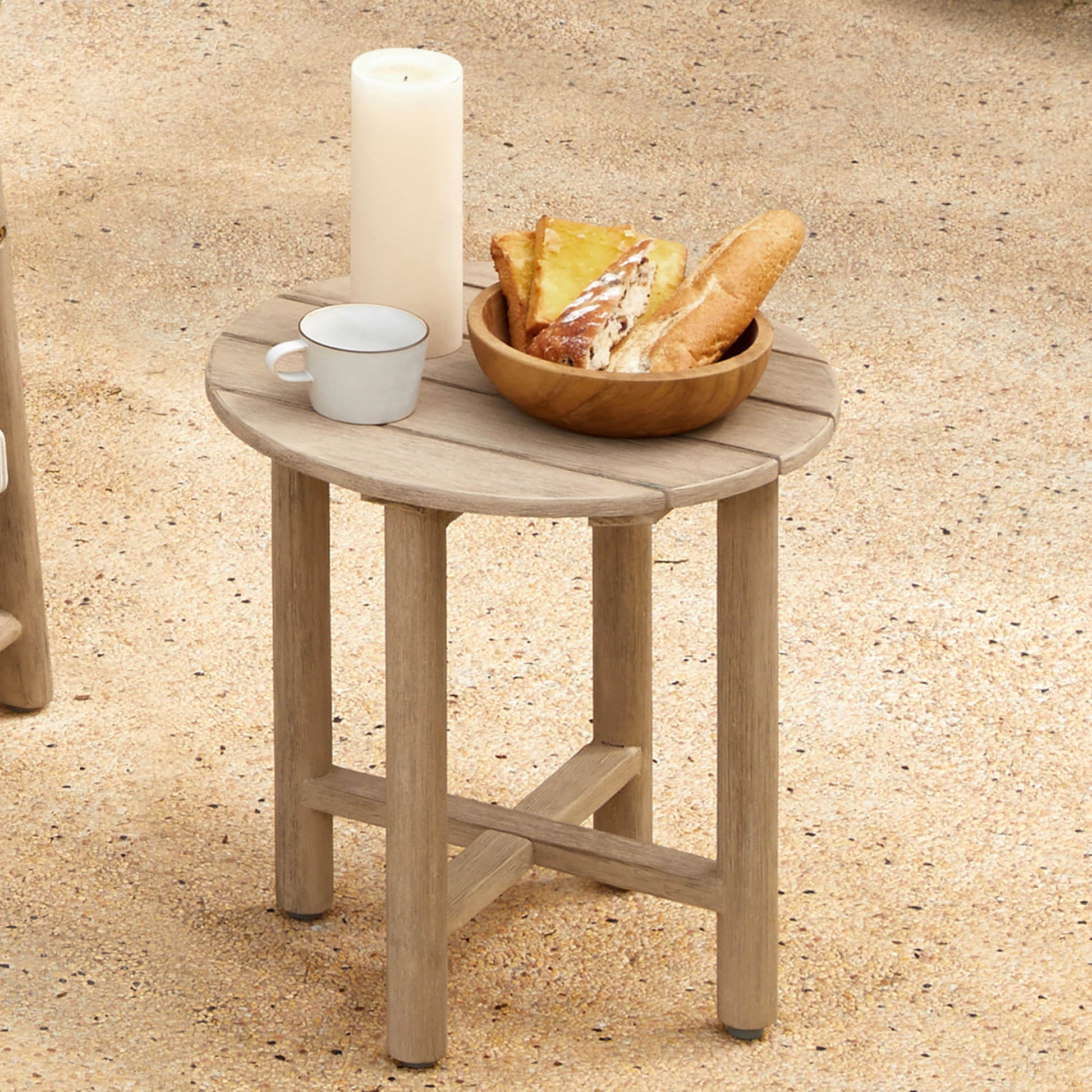 A Sienna Modern Acacia Wood Woven Outdoor Bistro Set table with a candle, mug, and bowl of bread on a sandy surface.