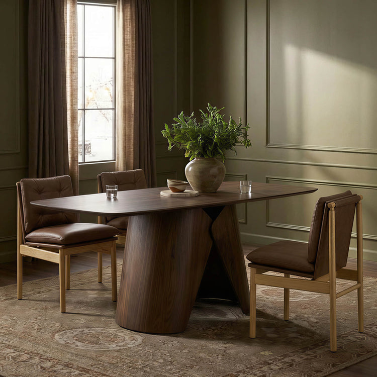 Elowen sculptural wood base dining table in walnut with angled pedestal and brown chairs.