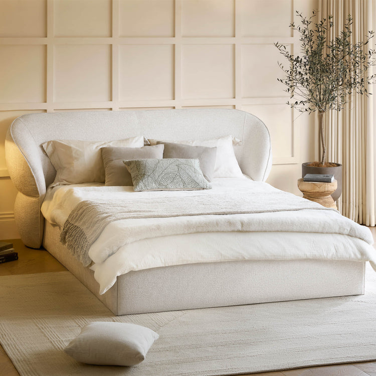 Beige upholstered bed with white bedding in a room with beige walls and a plant.