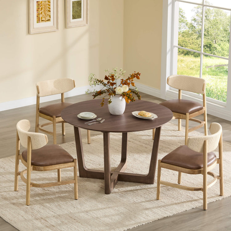 Rory round walnut dining table with a cross-base, paired with four wood and woven dining chairs with brown leather seats.