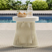 A white side table with a woven base, topped with a basket of oranges and a glass bottle, poolside.