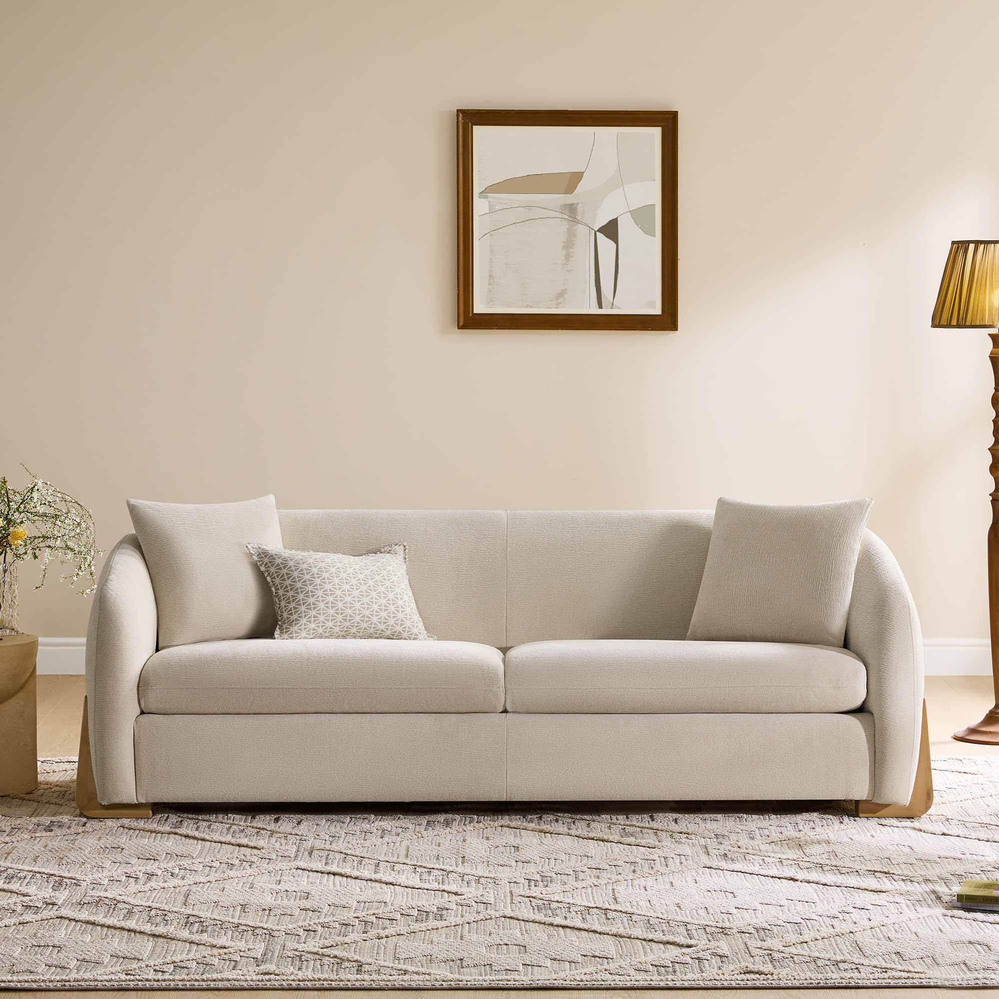  A cozy cream sofa with rounded armrests and wooden legs, set in a warmly lit living room with abstract art on the wall and a patterned rug on the floor.