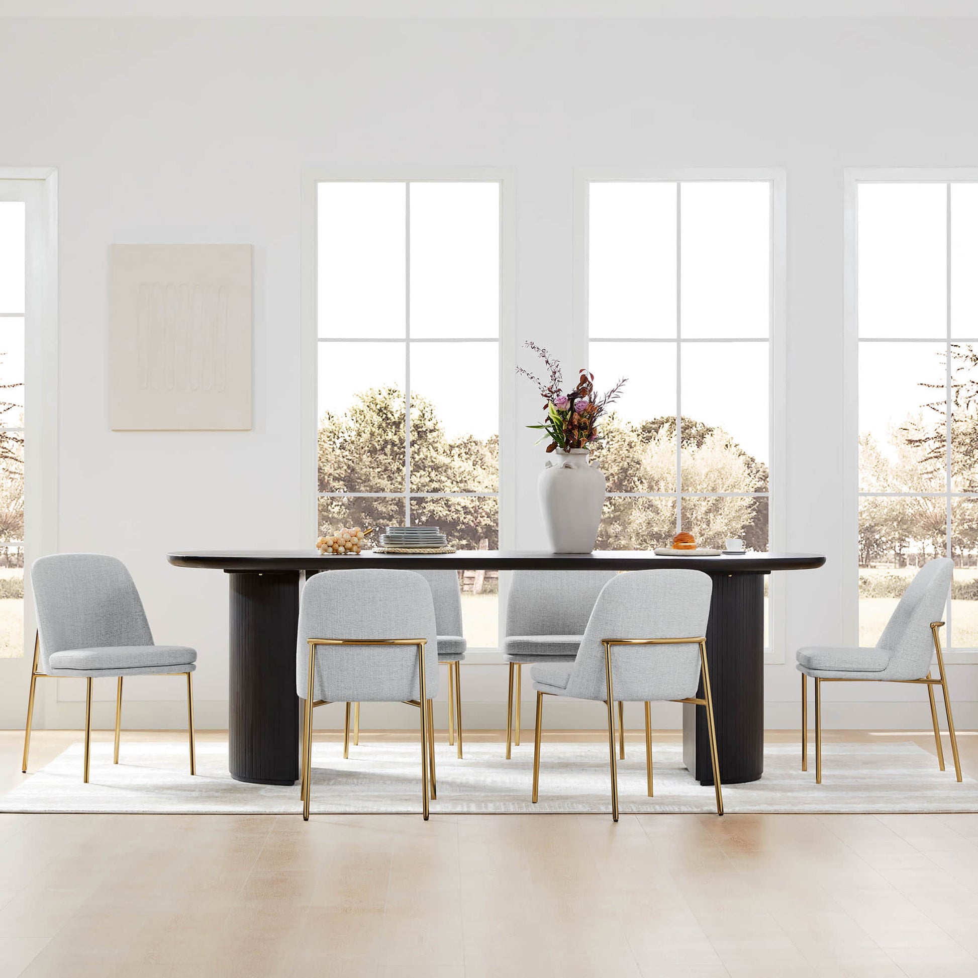 Golda Dining Chair, light gray fabric with gold legs, in a bright dining room with large windows.