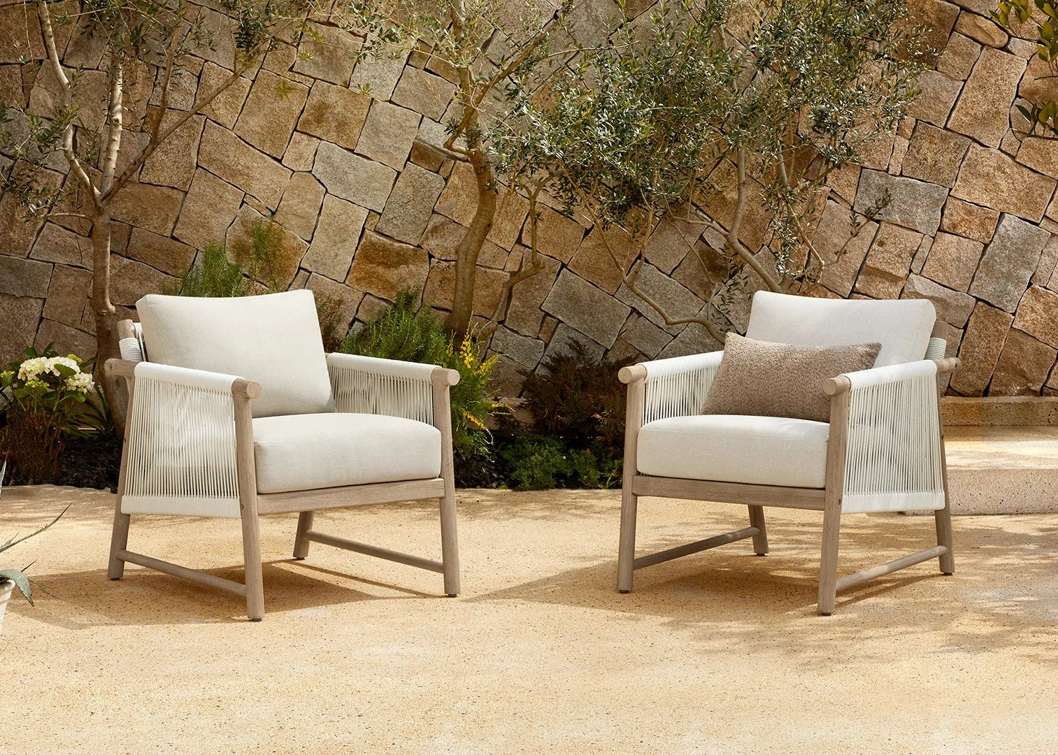 Two outdoor armchairs with light beige woven rope seats and backs, set on a stone patio in front of a rustic stone wall with plants.
